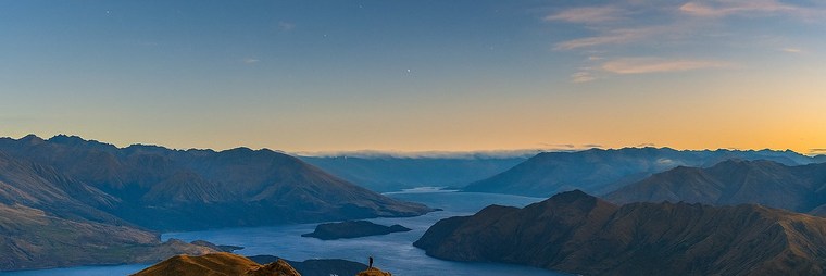 A solitary figure stands on a ridgeline above Lake Wanaka at dawn, looking out across the mountains of Aotearoa New Zealand.