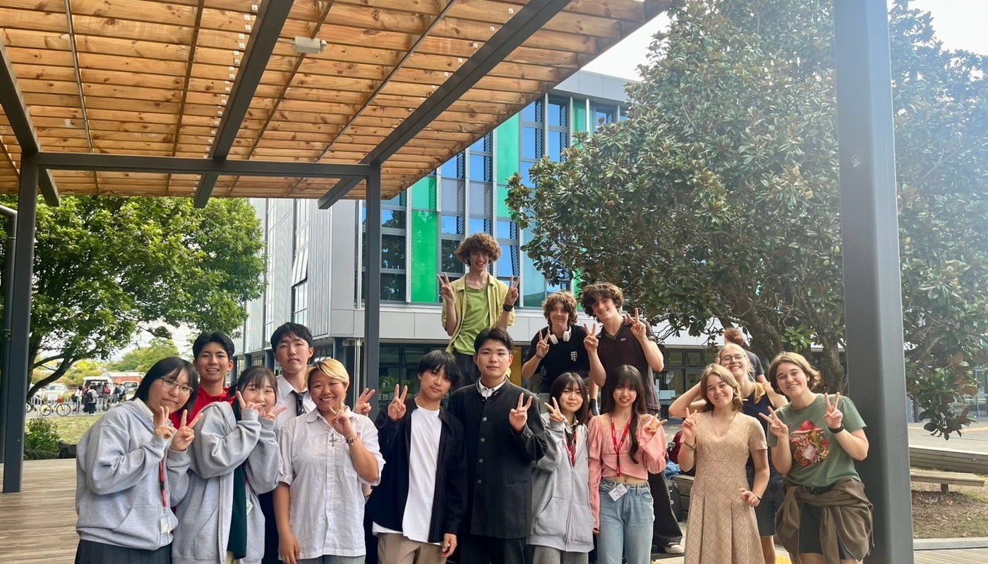 A group of international and New Zealand students gathered together outside a Kiwi school, smiling and making peace signs.