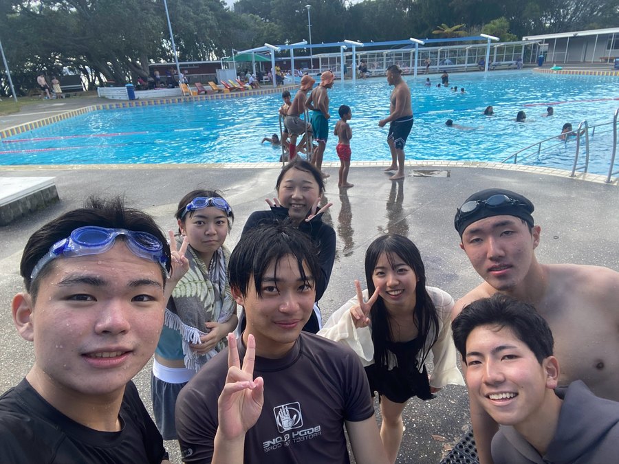 A group of students together at a Kiwi public swimming pool — laughing, peace signs, goggles up.