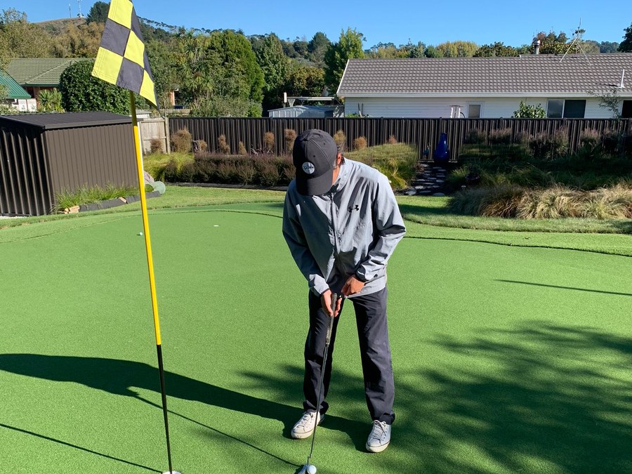 A golfer lining up a putt on a sunlit Kiwi practice green, mountains rising in the distance.