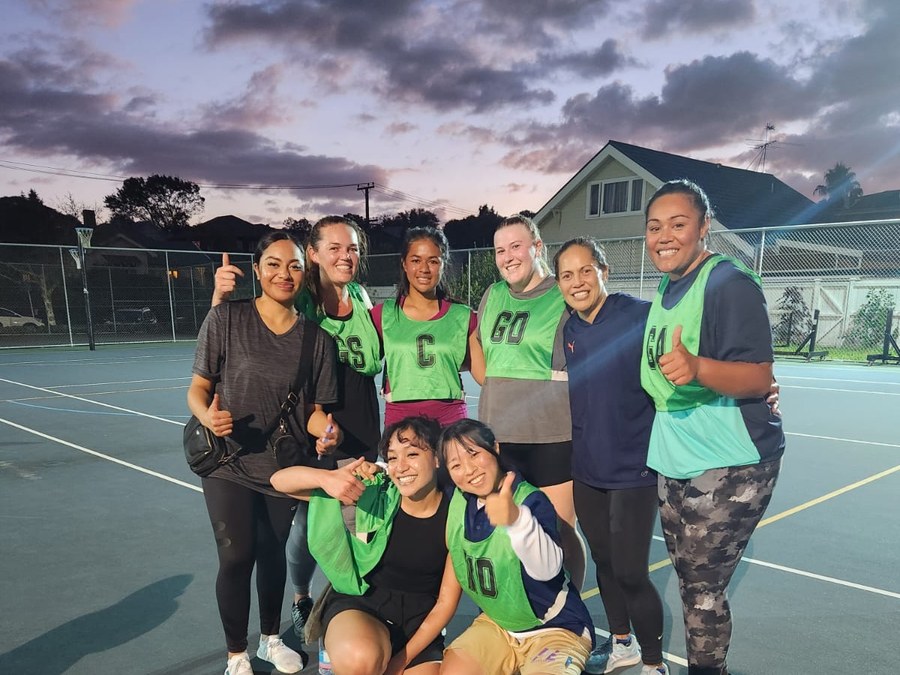 A community netball team gathered together on court at dusk after a game, smiling and laughing — the kind of belonging that good sport builds.