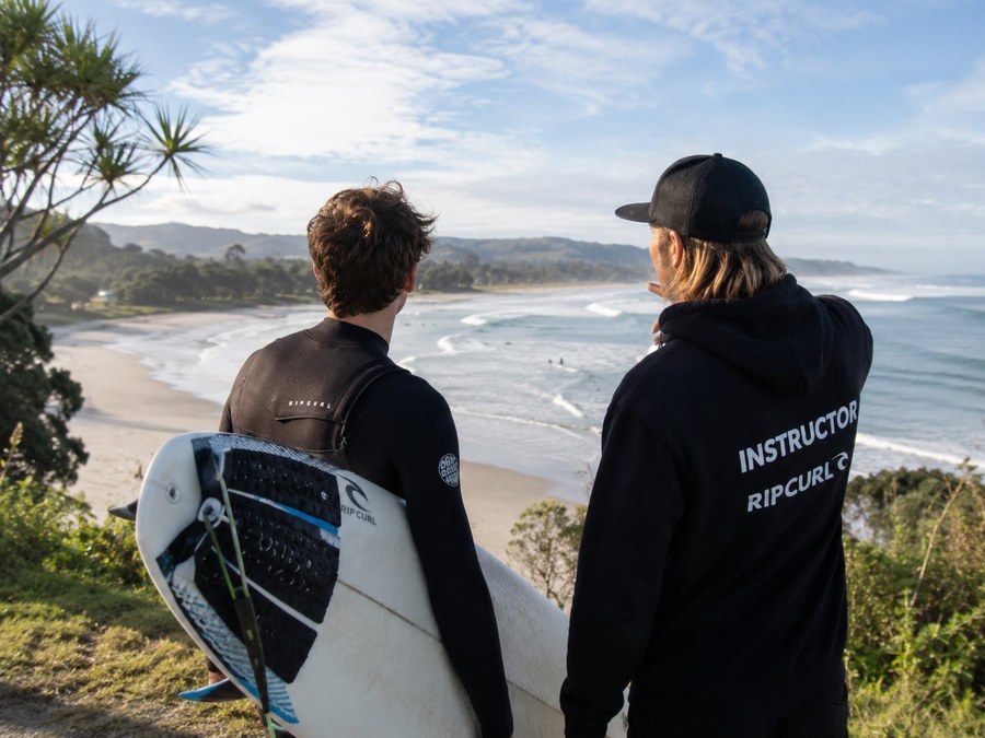 A young surfer and an instructor standing on a Kiwi headland, watching the waves break together.